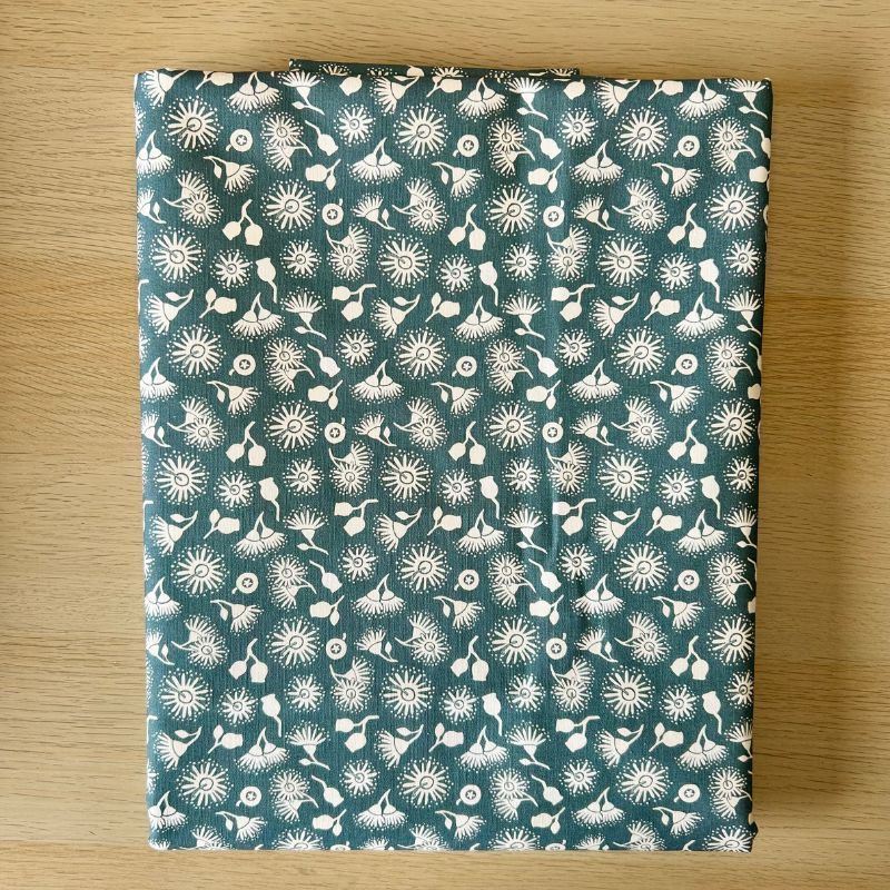 Cotton canvas, featuring a white floral and dandelion-like pattern on a dark teal-green background, laid flat on a light wood surface.