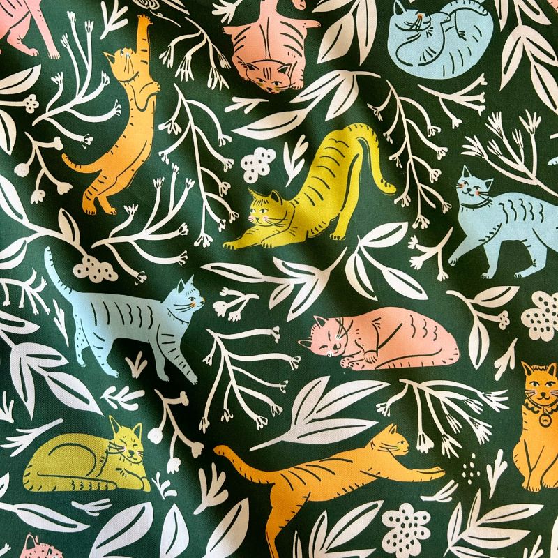 Dark green fabric featuring a playful pattern of illustrated cats in orange, yellow, light blue, and pink, interspersed with white leafy branches and floral motifs.