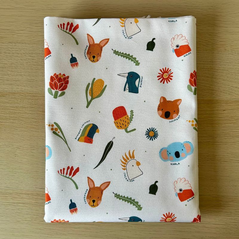 A folded white fabric cloth featuring a colorful Australian-themed animal and floral print, laid flat on a light wood surface.