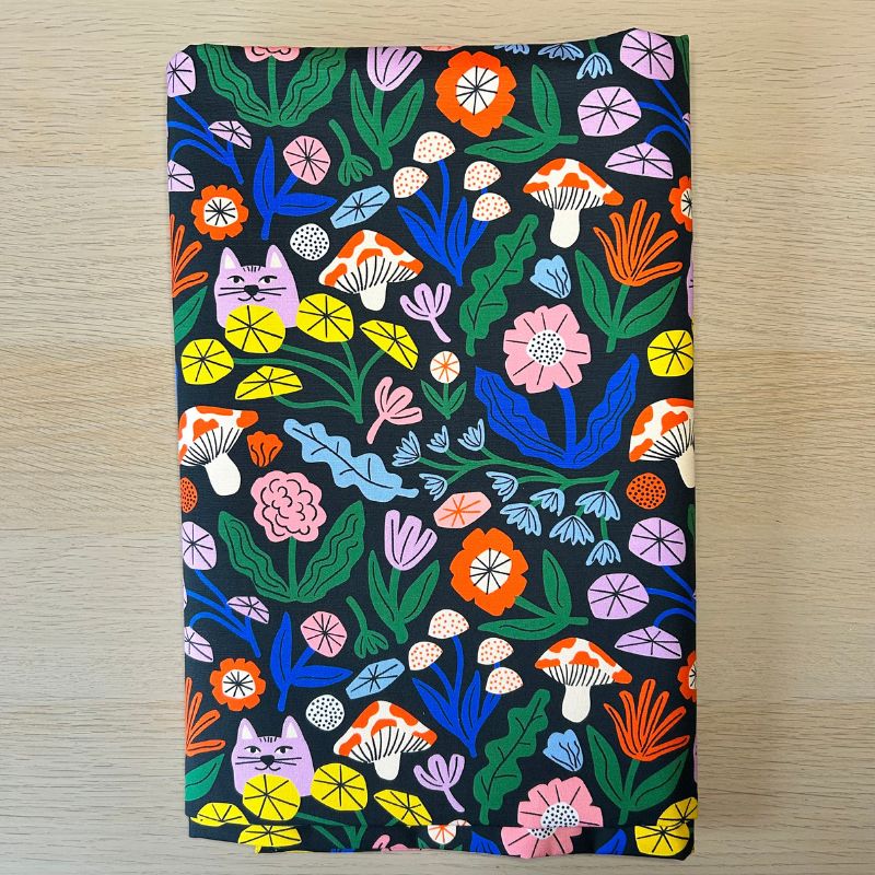 A folded piece of fabric with a colorful, whimsical pattern featuring a purple cat, mushrooms, flowers, and leaves on a black background, displayed flat on a light wood surface.