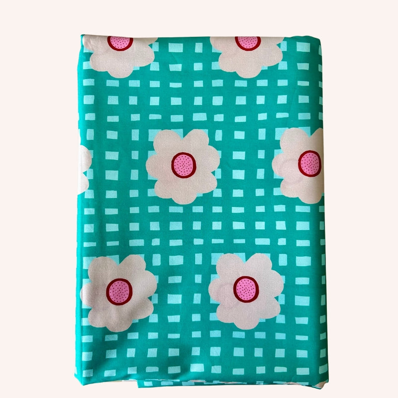 A folded piece of fabric with a teal and white gingham pattern, decorated with large white flowers that have pink centers, shown against a light beige background.