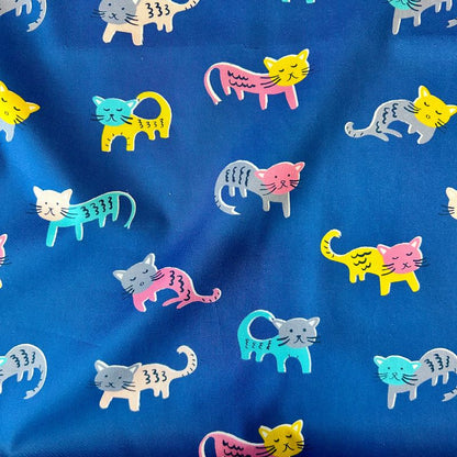 A close-up of blue fabric printed with a repeating pattern of stylized walking cats in pink, yellow, teal, gray, and white.