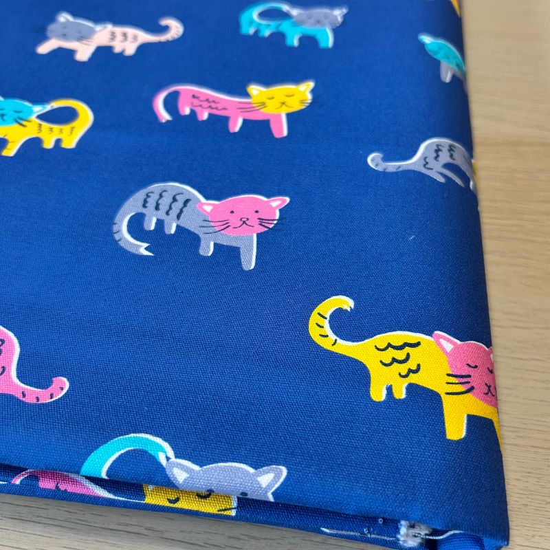 A close-up of folded blue fabric printed with colorful cartoon cats in pink, yellow, gray, and turquoise, laid on a light wood surface.