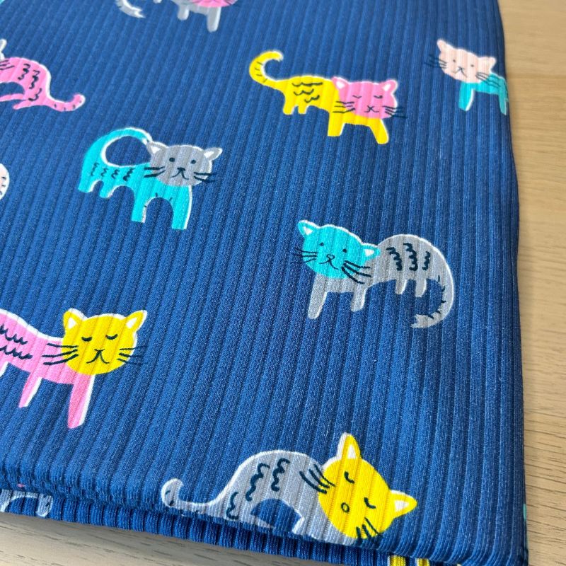 A close-up of a blue ribbed fabric featuring a playful pattern of cartoon cats in pink, yellow, teal, and gray, laid flat on a light wood surface.