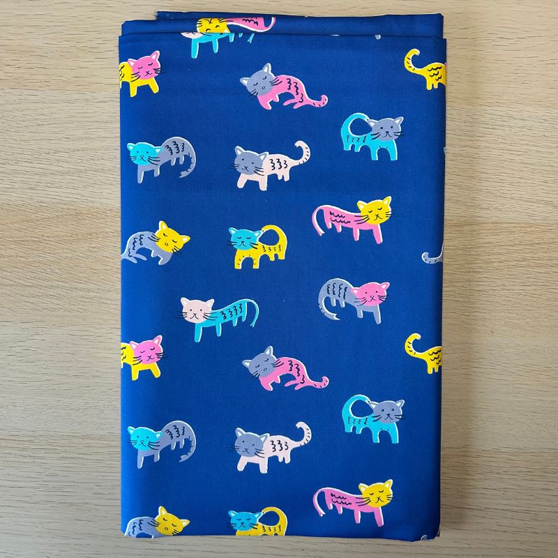 A folded piece of blue fabric with a repeating pattern of colorful cartoon cats in pink, yellow, blue, gray, and white, displayed flat on a light wood surface.