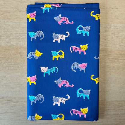 A folded piece of blue fabric with a repeating pattern of colorful cartoon cats in pink, yellow, blue, gray, and white, displayed flat on a light wood surface.