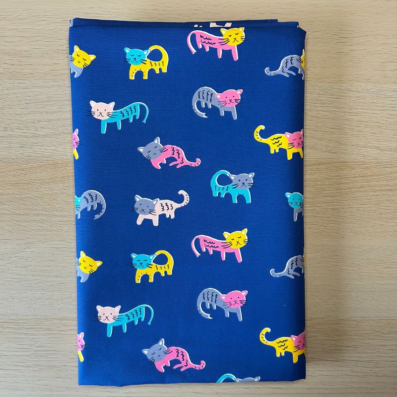 A folded piece of royal blue fabric with a playful pattern of colorful cartoon cats in pink, yellow, teal, and gray, displayed flat on a light wood surface.