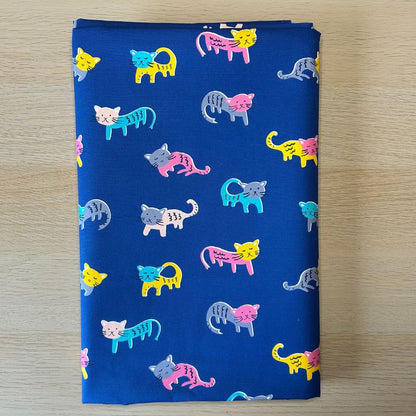 A folded piece of royal blue fabric with a playful pattern of colorful cartoon cats in pink, yellow, teal, and gray, displayed flat on a light wood surface.