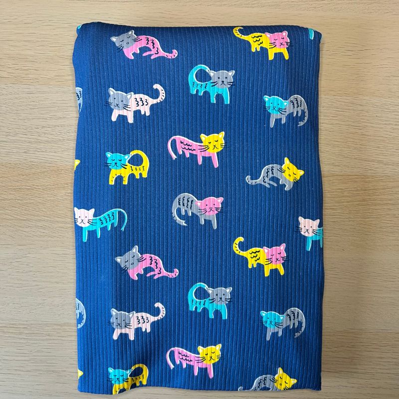 A folded piece of blue ribbed fabric featuring a colorful pattern of cartoon cats in pink, yellow, teal, and gray, laid flat on a light wood surface.