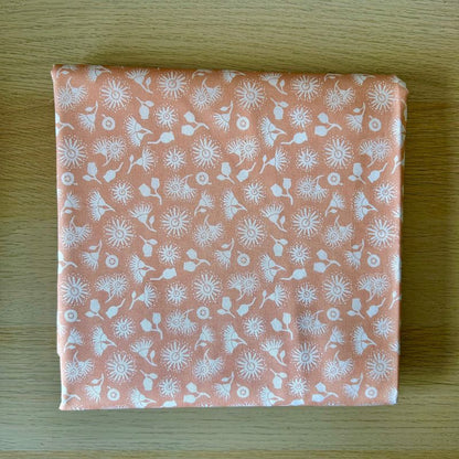 A square piece of peach-colored fabric with a white dandelion pattern, folded neatly on a light wood grain surface.