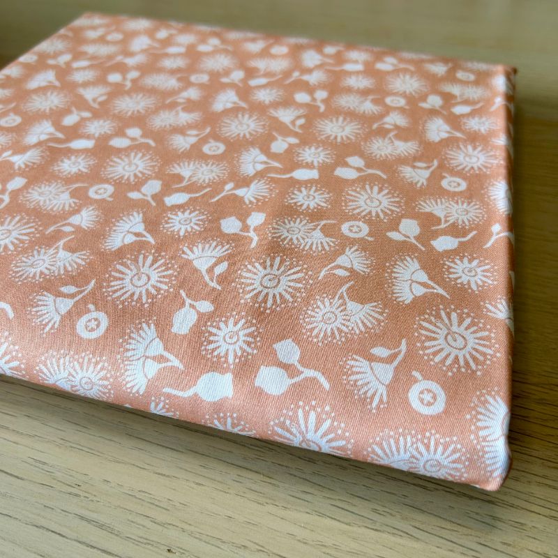 A square fabric item, likely a small bag or pouch, featuring a white dandelion and floral pattern on a dusty peach background, resting on a light wood surface.