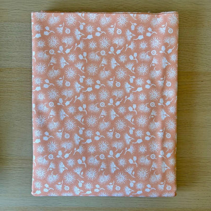A folded piece of peach-colored fabric with a white all-over floral and botanical pattern, laid flat on a light wood surface.