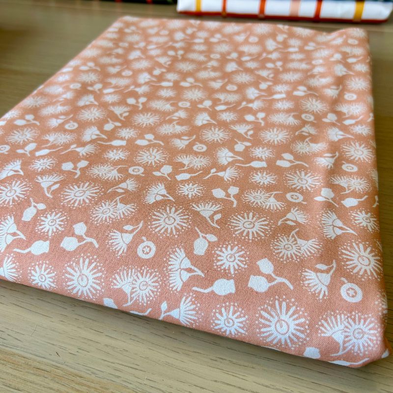 A folded piece of peach-colored fabric with a white hand-drawn style floral and leaf pattern, resting on a light wood surface.