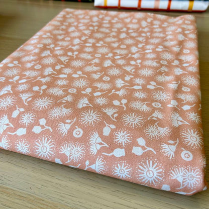 A folded piece of peach-colored fabric with a white hand-drawn style floral and leaf pattern, resting on a light wood surface.