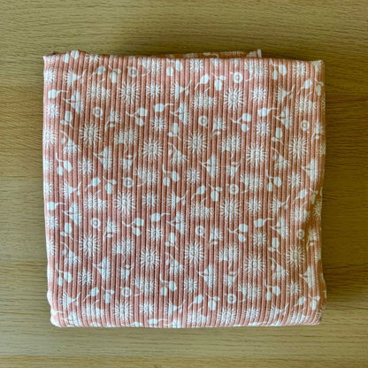 A folded piece of textured pink fabric with a white floral and botanical pattern, displayed flat on a light wood surface.
