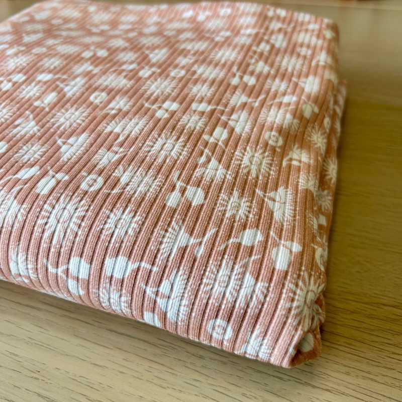 A folded piece of ribbed fabric in a dusty terracotta pink color with a white repeating botanical and abstract pattern, displayed on a light wood surface.
