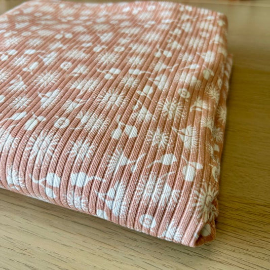 A folded piece of ribbed fabric in a dusty terracotta pink color with a white repeating botanical and abstract pattern, displayed on a light wood surface.