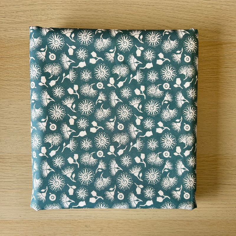 A square fabric item,  featuring a white dandelion and floral pattern on a teal blue-green background, displayed flat on a light wood surface.