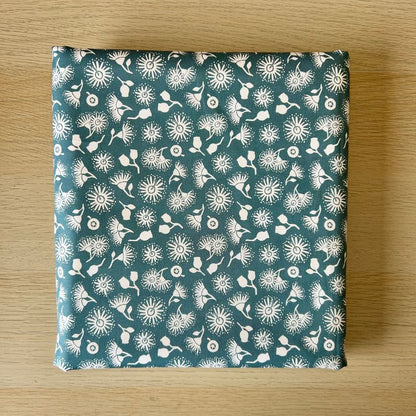 A square fabric item,  featuring a white dandelion and floral pattern on a teal blue-green background, displayed flat on a light wood surface.