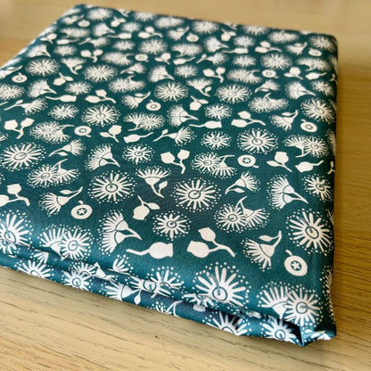 A folded piece of fabric with a dark teal green background and a white repeating floral and abstract pattern, placed on a light wood surface.
