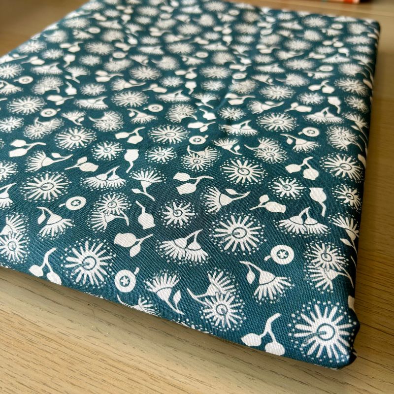 A folded piece of fabric with a dark teal blue background and a white repeating floral and abstract botanical pattern, displayed on a light wood surface.