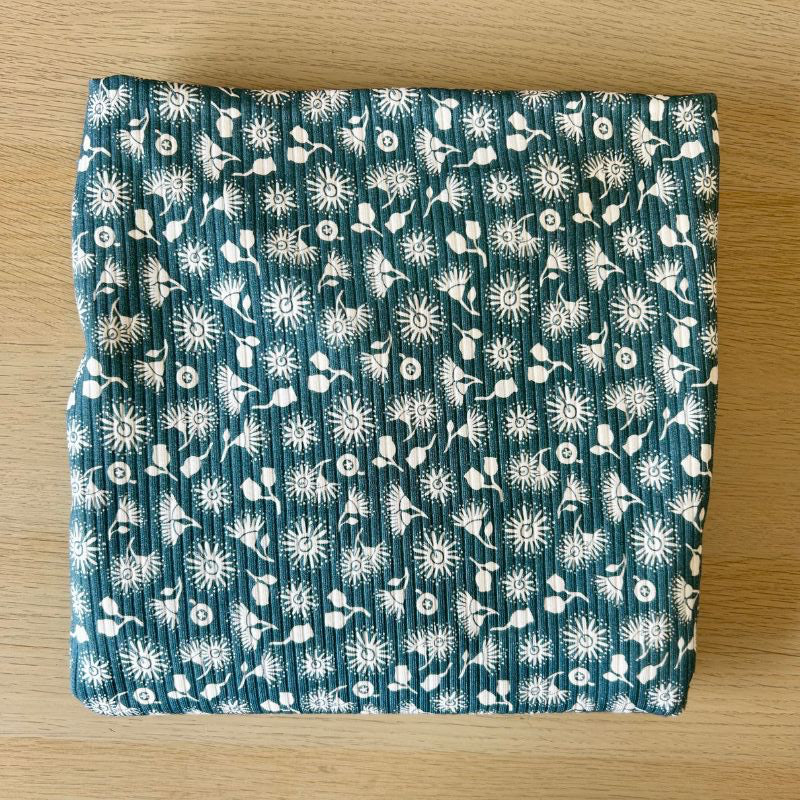 A folded piece of ribbed knit fabric with a white dandelion and floral pattern on a teal blue background, placed on a light wood surface.