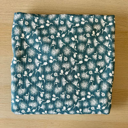 A folded piece of ribbed knit fabric with a white dandelion and floral pattern on a teal blue background, placed on a light wood surface.