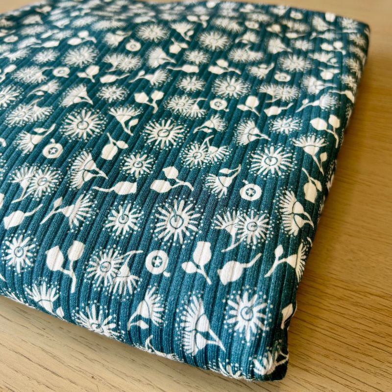 Ribbed knit fabricwith a white floral and botanical block print on a dark teal-green background, resting on a light wood surface.