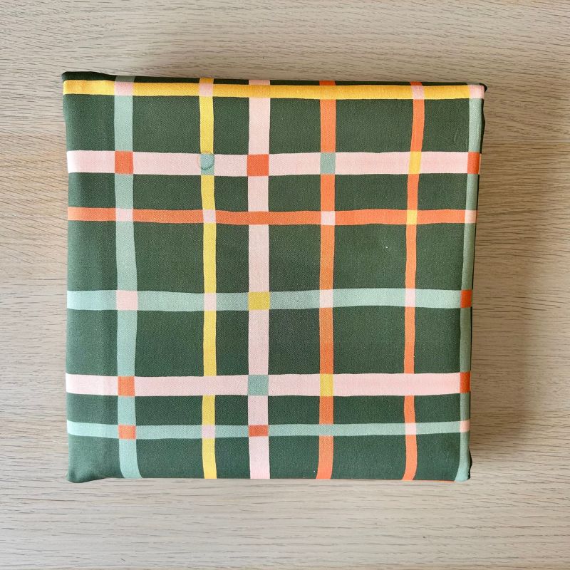 Cotton fabric, featuring a plaid pattern in dark green with intersecting stripes of orange, yellow, light pink, and pale blue, laid flat on a light wood surface.