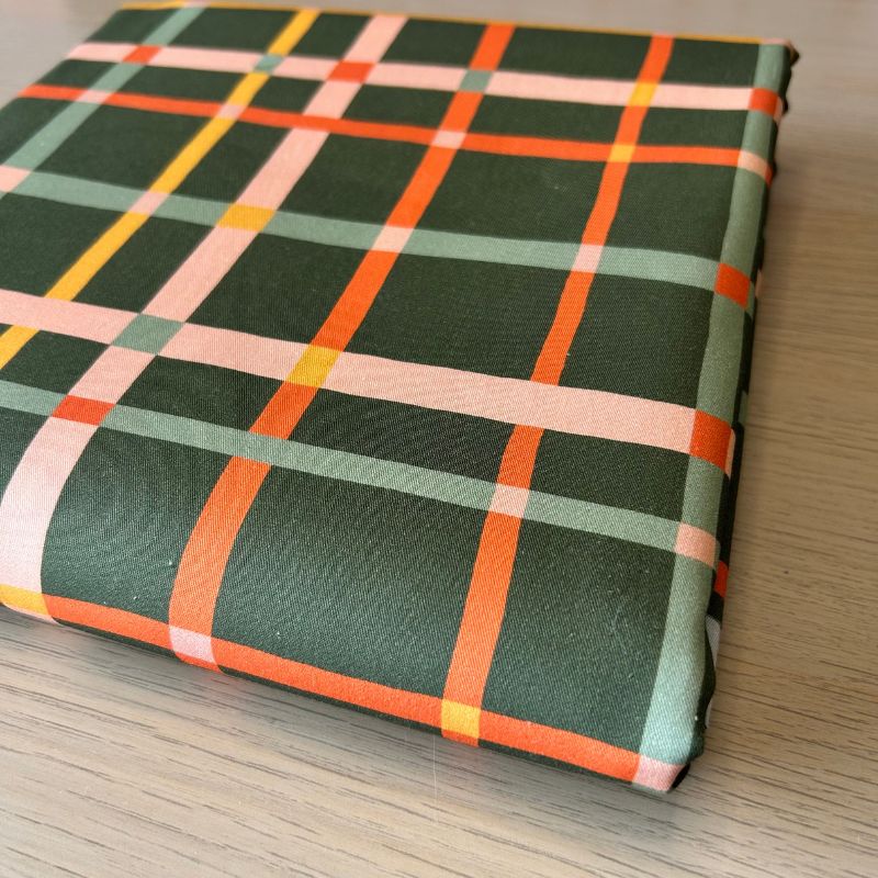 Cotton fabric with a plaid pattern in dark green, orange, light pink, and pale yellow, resting on a light wood surface.