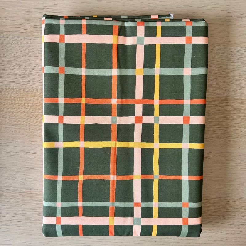 Cotton canvas featuring a dark green plaid pattern with intersecting lines in orange, yellow, light blue, and pale pink, laid flat on a light wood surface.