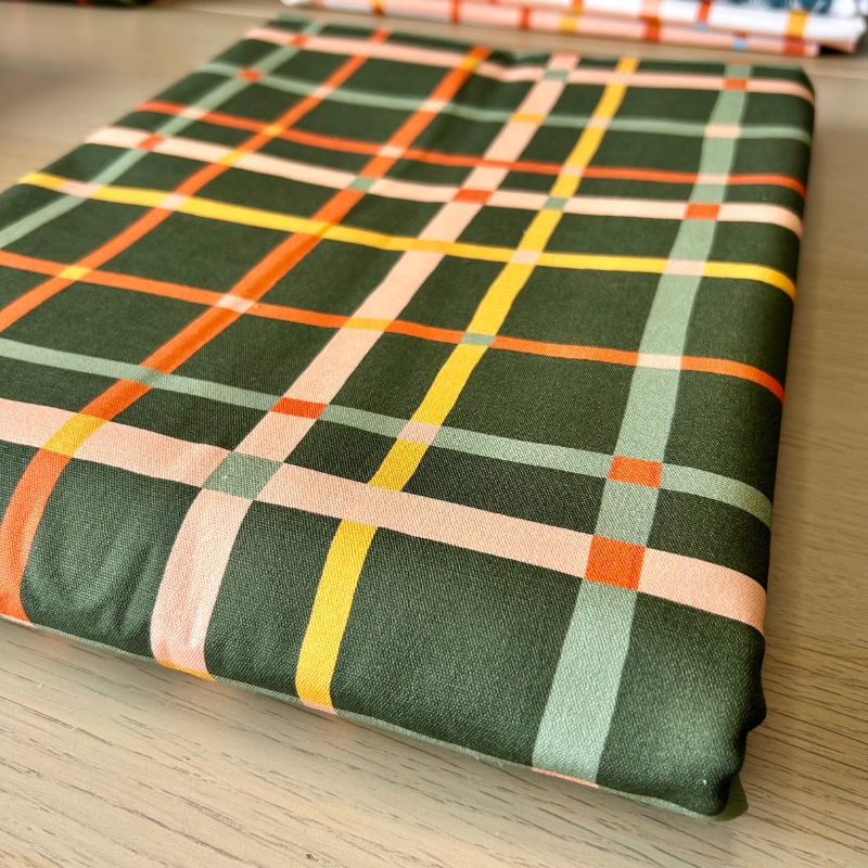 A folded piece of fabric with a green plaid pattern featuring intersecting stripes of orange, yellow, light green, and peach, displayed on a light wood surface.