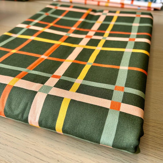 A folded piece of fabric with a green plaid pattern featuring intersecting stripes of orange, yellow, light green, and peach, displayed on a light wood surface.