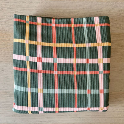 Ribbed fabric  with a plaid pattern in dark green, pink, orange, and light blue, laid flat on a light wood surface.