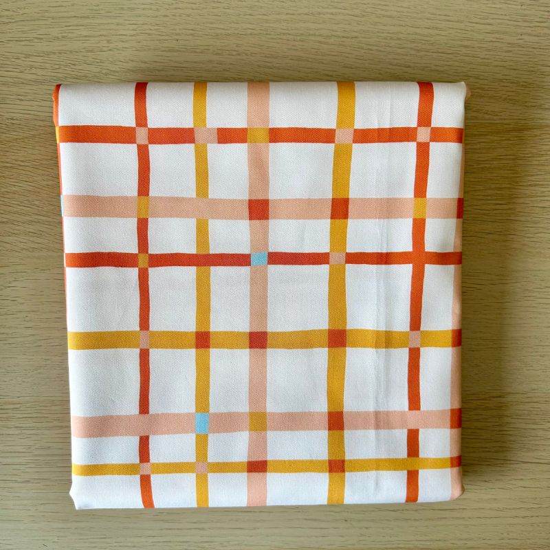 A folded piece of white fabric with a geometric plaid pattern in orange, yellow, and peach, placed on a light wood surface.