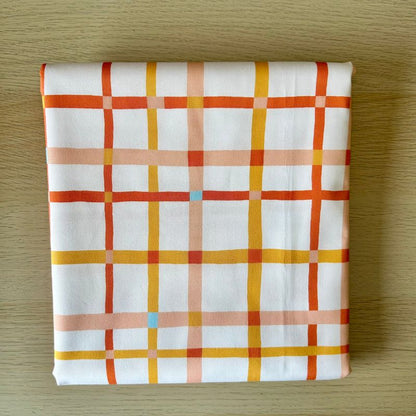 A folded piece of white fabric with a geometric plaid pattern in orange, yellow, and peach, placed on a light wood surface.