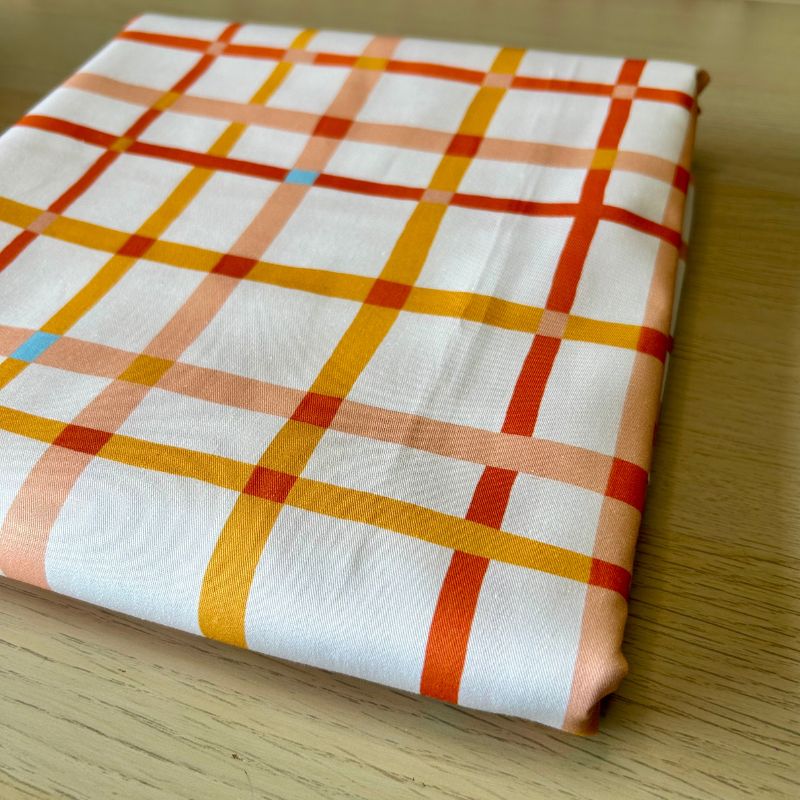 A folded white fabric with a plaid pattern in orange, yellow, and peach, resting on a light wood surface.