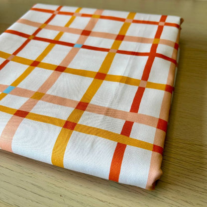 A folded white fabric with a plaid pattern in orange, yellow, and peach, resting on a light wood surface.