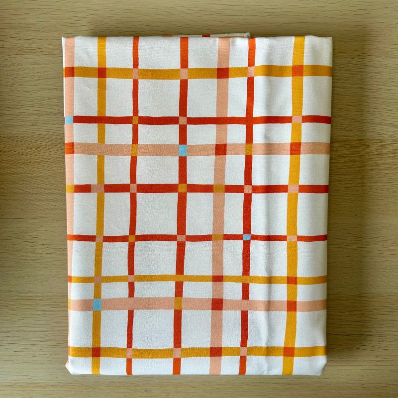 White fabric featuring a plaid pattern with intersecting orange, red, and peach lines, laid flat on a light wood surface.