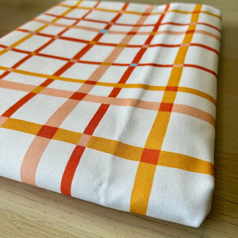 White cotton canvas featuring a warm plaid pattern with intersecting orange, red, and yellow stripes, displayed on a light wood surface.