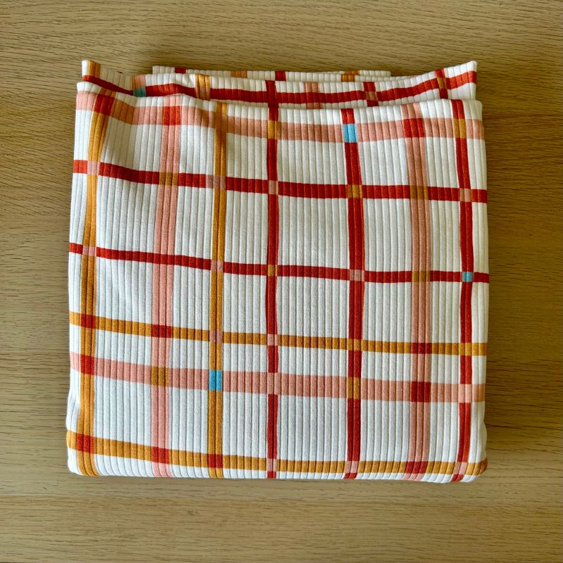 White ribbed fabric with a red, orange, and pink plaid pattern, laid flat on a light brown wooden surface.