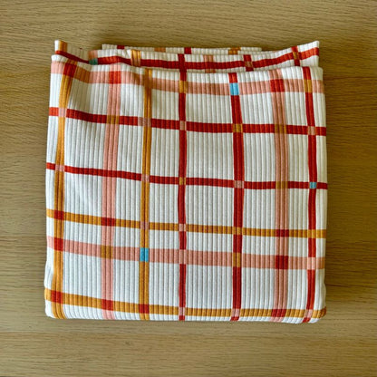 White ribbed fabric with a red, orange, and pink plaid pattern, laid flat on a light brown wooden surface.