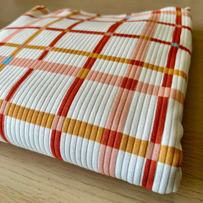 A folded piece of white ribbed fabric with a warm-toned plaid pattern in orange, red, and peach, resting on a light wood surface.
