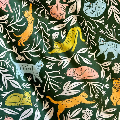 Dark green fabric featuring a playful pattern of illustrated cats in orange, yellow, light blue, and pink, interspersed with white leafy branches and floral motifs.