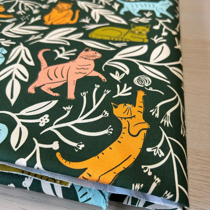 A folded piece of dark green fabric with a whimsical pattern of stylized orange, yellow, and blue cats among white leafy vines, shown on a light wood surface.