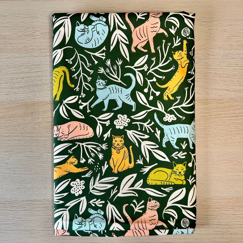 A piece of folded fabric  featuring a playful pattern of illustrated cats in yellow, light blue, and pink on a dark green background with white leaves. 