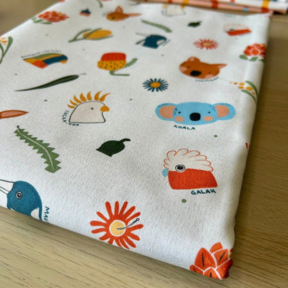 White fabric printed with colorful cartoon animals and objects, including a koala, galah, bird, flower, and leaf, displayed on a light wood surface.