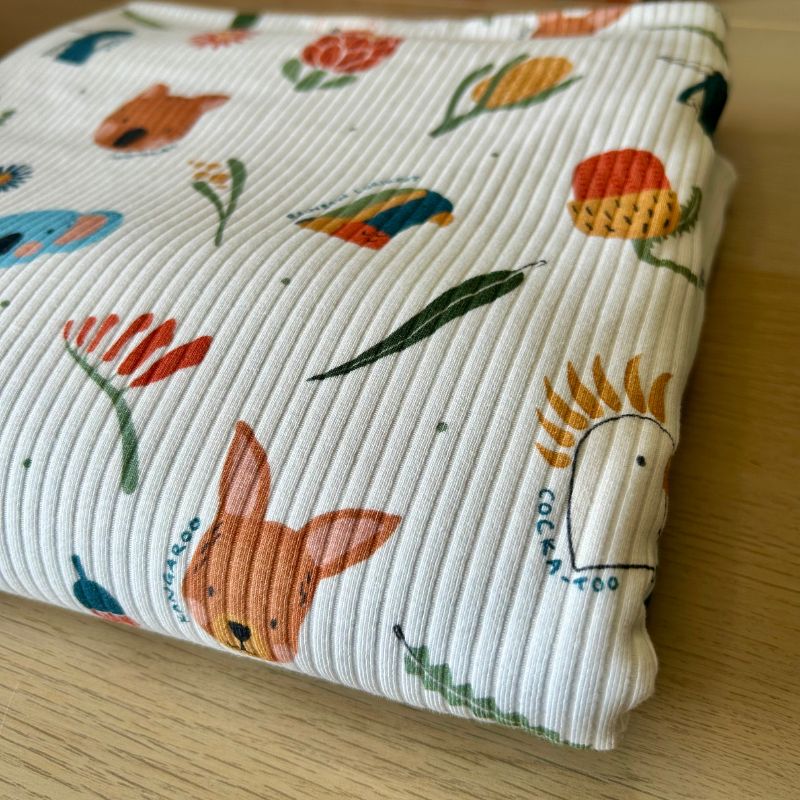 A folded white ribbed fabric item, likely a baby blanket or muslin cloth, featuring a colorful print of cartoon animals and flowers, resting on a light wood surface.