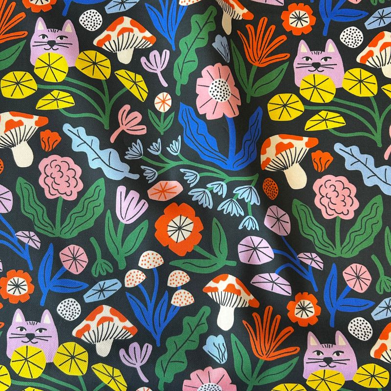 A close-up of a black fabric with a colorful, playful pattern featuring cartoon cats, mushrooms, flowers, and leaves in pink, orange, yellow, blue, green, and purple.