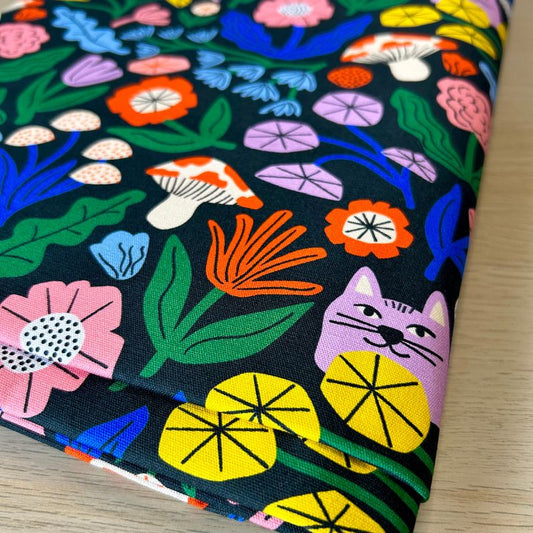 Folded fabric with a colorful floral and mushroom print on a black background, photographed on a light wood surface.